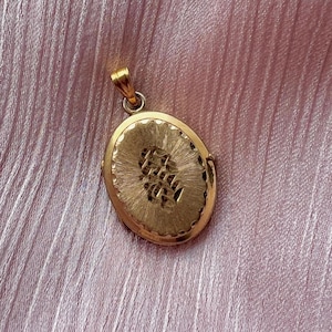 May include: An oval gold-toned pendant featuring a textured, sunburst-style engraved pattern on the front surface. The pendant includes a small, integrated bail at the top for attachment to a chain, showcasing a polished, metallic finish throughout.