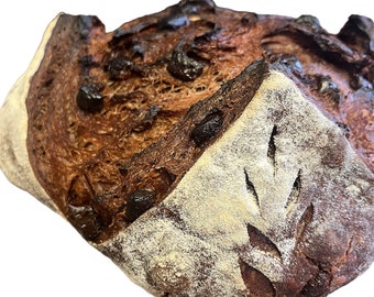 May include: A dark brown sourdough bread loaf with a light brown crust and a leaf design scored into the top. The bread has a rustic appearance and is speckled with dark chocolate chips.