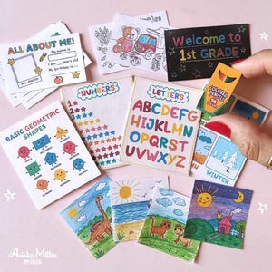 Miniature school supplies set including tiny flashcards, alphabet and numbers charts, basic geometric shapes guide, colorful drawings, and a mini colored pencils box, displayed on a pink background with a hand holding one item. Perfect for dollhouses, crafts, or back-to-school decor by Aninka Miller Minis