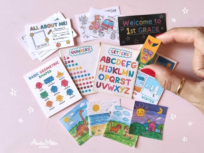 Miniature school supplies set including tiny flashcards, alphabet and numbers charts, basic geometric shapes guide, colorful drawings, and a mini colored pencils box, displayed on a pink background with a hand holding one item. Perfect for dollhouses, crafts, or back-to-school decor by Aninka Miller Minis