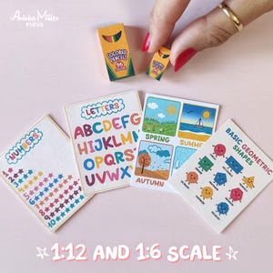 Miniature school printables and supplies in 1:12 and 1:6 scale. Tiny educational posters with alphabet letters, numbers, seasons, and geometric shapes, plus miniature colored pencil boxes, shown with a hand for scale. Perfect for dollhouse classrooms and miniature crafts by Aninka Miller Minis