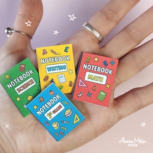 Set of colorful printable miniature school notebooks for dollhouses or crafts, including math, science, writing, and 1st grade notebooks. DIY mini books held in hand, perfect for dollhouse accessories and handmade miniatures. By Aninka Miller Minis