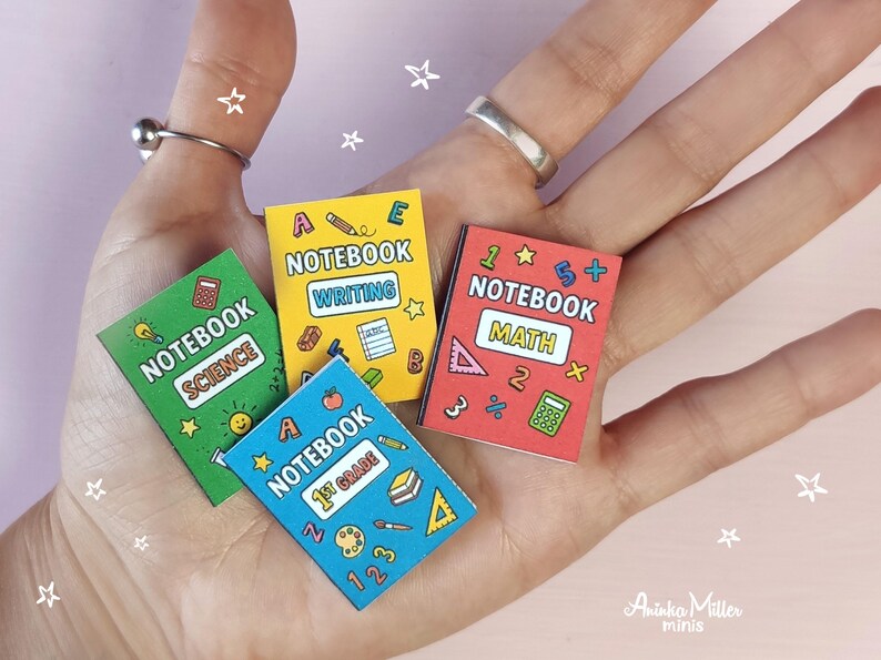 Set of colorful printable miniature school notebooks for dollhouses or crafts, including math, science, writing, and 1st grade notebooks. DIY mini books held in hand, perfect for dollhouse accessories and handmade miniatures. By Aninka Miller Minis