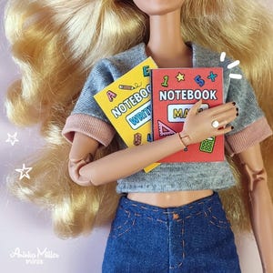 Printable miniature school notebooks for dollhouses or crafts, shown with a fashion doll holding colorful math and writing notebooks. DIY mini books, perfect for dollhouse accessories, school-themed miniatures, and handmade crafts. By Aninka Miller Minis