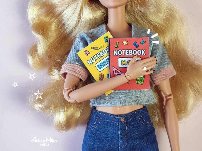 Printable miniature school notebooks for dollhouses or crafts, shown with a fashion doll holding colorful math and writing notebooks. DIY mini books, perfect for dollhouse accessories, school-themed miniatures, and handmade crafts. By Aninka Miller Minis