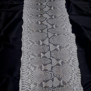 Peut inclure: Motif de peau de serpent dans des tons de gris et de blanc, avec un motif central en losange et un aspect écailleux texturé. La peau de serpent est présentée sur un fond noir.
