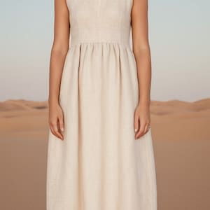 Può includere: Una donna indossa un abito di lino beige chiaro con scollo rotondo e vita arricciata. È in piedi in un ambiente desertico.