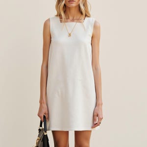 Può includere: Un abito bianco senza maniche, corto, con scollo quadrato e una collana a catena dorata. L'abito è realizzato in un tessuto leggero e ha una vestibilità rilassata.