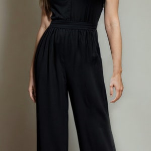 May include: A woman wearing a black sleeveless jumpsuit with a V-neckline and a button closure. The jumpsuit has a wide leg and a fitted bodice.