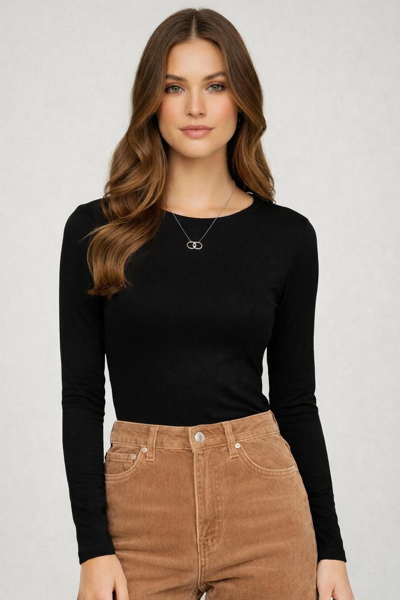 May include: A black long-sleeve shirt paired with brown corduroy pants. A silver necklace with a double-circle pendant is visible. The model is wearing a casual outfit.