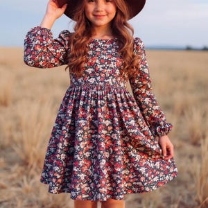 May include: A young person wearing a brown hat and a long-sleeved dress with a floral pattern in red, white, and pink on a navy blue background. The dress has a flared skirt and is paired with brown boots.