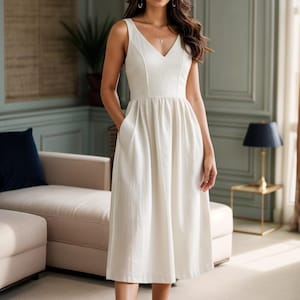May include: A white sleeveless midi dress with a V-neckline and a fitted bodice. The dress has a flowy skirt and pockets on the sides. The model is wearing the dress with sandals.