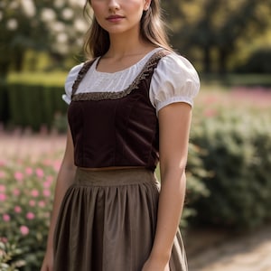May include: A woman wearing a brown velvet bodice with a white blouse and a brown skirt. She is standing in a garden with flowers.