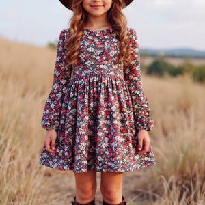 May include: A young person wearing a brown hat, a floral print dress with long sleeves, and brown boots. The dress features a dark background with red and white flowers. The person is standing in a field of tall grass.