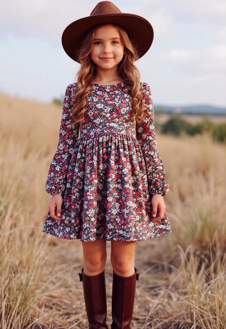 May include: A young person wearing a brown hat, a floral print dress with long sleeves, and brown boots. The dress features a dark background with red and white flowers. The person is standing in a field of tall grass.