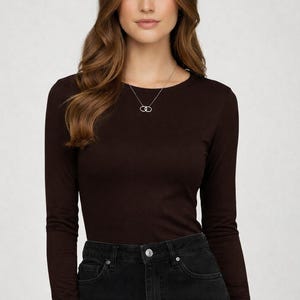 May include: A woman wearing a long-sleeved, dark brown shirt and black jeans. A silver necklace with a double-circle pendant is visible. The shirt has a crew neck and a fitted silhouette.