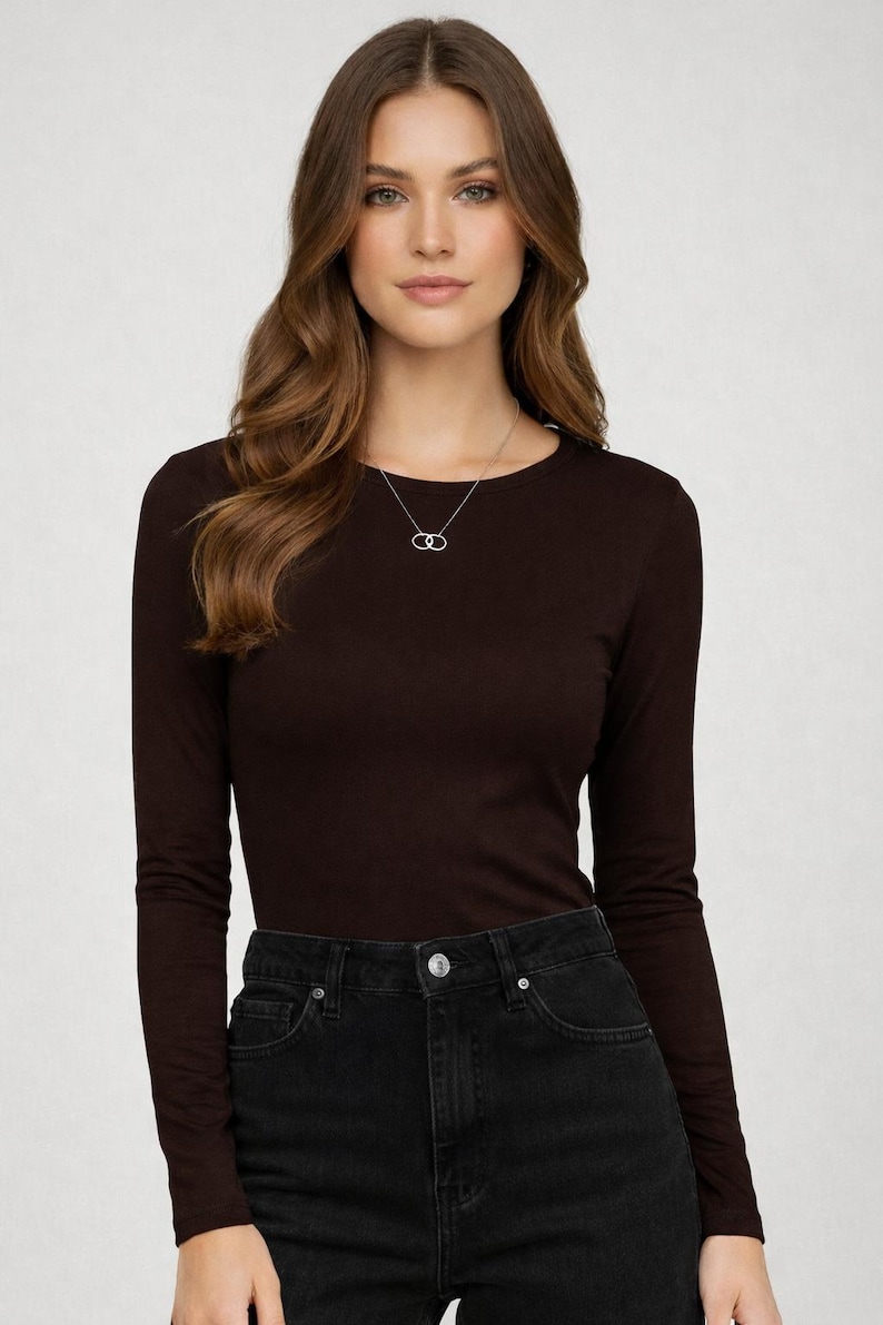 May include: A woman wearing a long-sleeved, dark brown shirt and black jeans. A silver necklace with a double-circle pendant is visible. The shirt has a crew neck and a fitted silhouette.