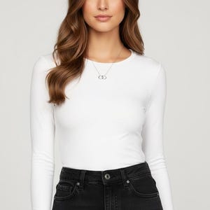 May include: A white long-sleeve shirt paired with black denim jeans. A silver necklace with two interlocking circles is visible. The model has long, wavy brown hair.