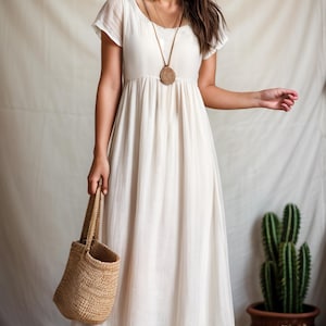 Puede incluir: Un vestido blanco maxi con escote cuadrado y mangas cortas. El vestido tiene una cintura fruncida y una falda fluida. La modelo lleva un collar marr&oacute;n con un gran colgante circular.