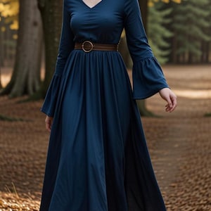 Può includere: Una donna che indossa un lungo abito blu scuro con una cintura marrone e stivali marroni. È in piedi in un bosco con foglie autunnali sul terreno.