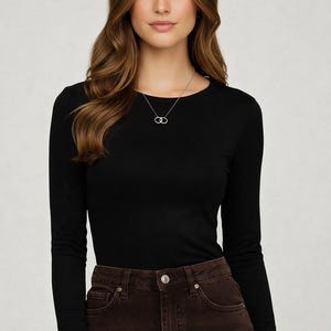 May include: A black long-sleeve shirt with a crew neck. The model is wearing a silver necklace with two interlocking circles. The shirt is paired with brown denim jeans.