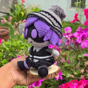 May include: A crocheted plush toy with purple hair, a black and white striped outfit, and a gray and white striped hat. The toy is sitting on a wooden base.