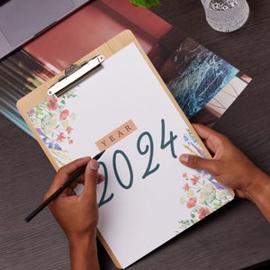 Peut inclure: Un presse-papiers blanc avec un motif floral et l'année 2024 écrite en bleu foncé. Le presse-papiers a un clip argenté et un cadre en bois brun.