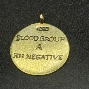 May include: A gold-coloured, circular pendant with a loop at the top. The pendant is engraved with the words "BLOOD GROUP A RH NEGATIVE". The pendant has a vintage appearance.