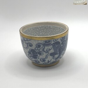 May include: A small, blue and white porcelain teacup with a gold rim and a dragon design. The cup has a crackled glaze and the bottom of the cup has the Chinese characters "年製" in blue.