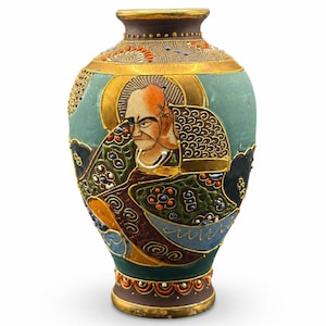 May include: A ceramic vase featuring a hand-painted portrait of a figure with a shaved head and prominent eyebrows. The vase is decorated with gold, teal, black, and orange accents, including intricate floral patterns and raised enamel details.