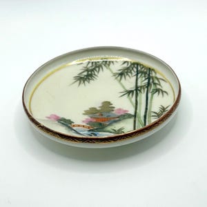 May include: Small, round decorative dish with an Asian landscape painting. The cream-coloured interior has a gold rim and a brown and gold patterned border. The landscape depicts bamboo, a bridge, and a pagoda.
