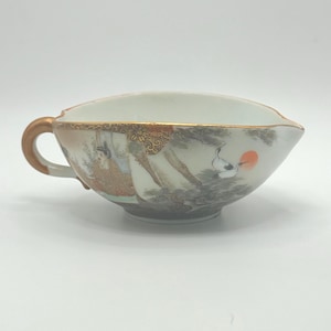 May include: A vintage Japanese porcelain teacup with a gold handle and rim. The cup features a painted scene of figures, birds, and a rising sun. The design includes shades of orange, brown, and grey.