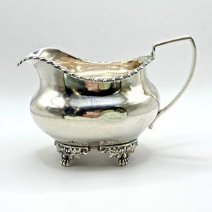 May include: A silver creamer with a curved handle and ornate, detailed feet. The rim of the creamer has a decorative edge. The creamer is reflective, showing the surrounding environment. The creamer is a classic piece for serving cream or milk.