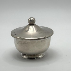 Op de afbeelding: Een zilveren suikerpot met een deksel en een ronde knop bovenop. De pot heeft een gladde, ronde vorm en een licht uitlopende basis.
