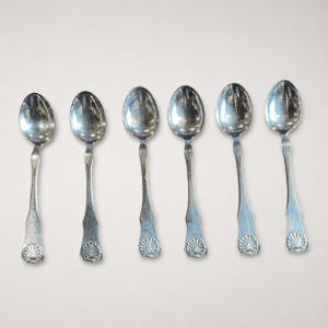 May include: A set of six silver-toned metal spoons arranged in a row. Each spoon features a polished oval bowl and a handle that terminates in a decorative, raised shell motif at the base. The metal surface has a reflective, metallic finish.