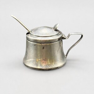May include: A small silver-coloured metal mustard pot with a hinged lid and a matching spoon. The container features a wide base, a curved handle on the side, and a small notch in the lid to accommodate the spoon handle. The surface shows a metallic finish.