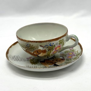 Könnte beinhalten: Ein weißes Porzellan-Tee-Set, bestehend aus einer Tasse und einer Untertasse. Die Tasse und die Untertasse sind mit braunen Vögeln, rosa und gelben Blumen und grünem Laub verziert. Der Rand der Tasse und der Untertasse hat einen goldenen Rand.