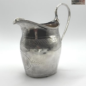 Puede incluir: Una jarra de plata con un asa curva y un pico. La jarra tiene grabados detallados alrededor de la parte superior e inferior, con un patrón texturizado en el medio. La jarra probablemente está hecha de plata de ley.