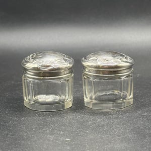 May include: Two small, clear glass jars with silver lids. The lids have a decorative design.