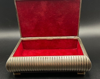 May include: A silver metal trinket box with a red velvet interior. The box has a ribbed design and sits on four small feet.