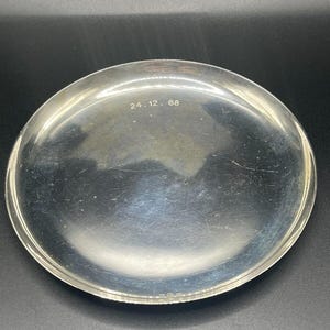 May include: A silver circular tray with a smooth surface and a raised rim. The tray has the inscription "24.12. 68" on the bottom.