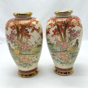 Puede incluir: Un par de jarrones de cerámica de color crema con detalles dorados. Los jarrones presentan un diseño de inspiración japonesa con hojas de arce doradas y rojas, flores rosas y faisanes dorados y azules.