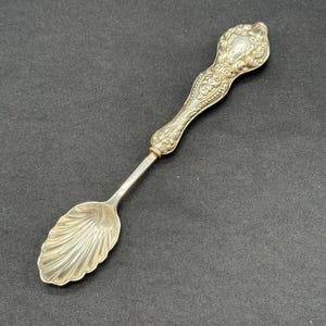 Puede incluir: Una cuchara de plata con un mango adornado y un cuenco en forma de concha. La cuchara mide aproximadamente 15 cm de largo.