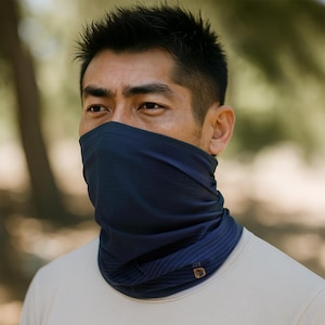 May include: A navy blue neck gaiter with subtle vertical stripes. The gaiter is worn around the neck and covers the lower face. A small, circular logo is visible on the lower right side. The background is blurred, suggesting an outdoor setting.