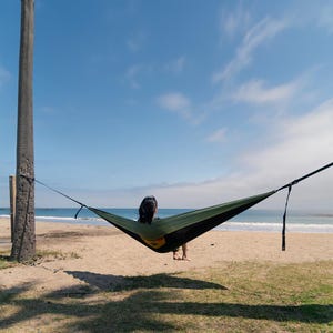 May include: A person relaxing in a green hammock suspended between a palm tree and a support on a sandy beach. The hammock is set against a backdrop of the ocean and a bright blue sky with scattered clouds.