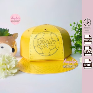 May include: A yellow papercraft baseball cap with a black outline of a soccer ball and the word "Dad" inside the ball. The cap is designed to be assembled.
