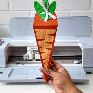Puede incluir: Una bolsa de papel para golosinas con forma de zanahoria con una parte superior de purpurina verde y una cinta blanca. La bolsa es naranja y tiene un patrón texturizado.