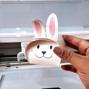 Peut inclure: Une boîte en papier blanche en forme de lapin avec des oreilles roses et un visage noir. La boîte est tenue dans une main.