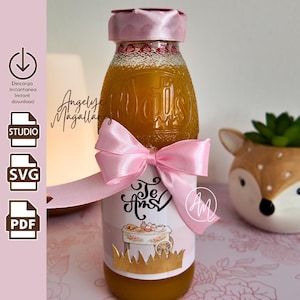 Puede incluir: Una botella de vidrio llena de líquido naranja, rematada con una tapa de cinta de satén rosa. Una etiqueta rosa y blanca presenta el texto Te Amo, un gráfico de un carrito de postres y un borde festoneado dorado. Un gran lazo de satén rosa al cuello.