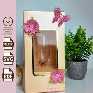 Puede incluir: Una copa de champán exhibida en una caja decorativa. La caja melocotón claro tiene una ventana con borde dorado, adornada con flores de papel rosa y una mariposa. El texto "Angelyc's Workshop" es visible.